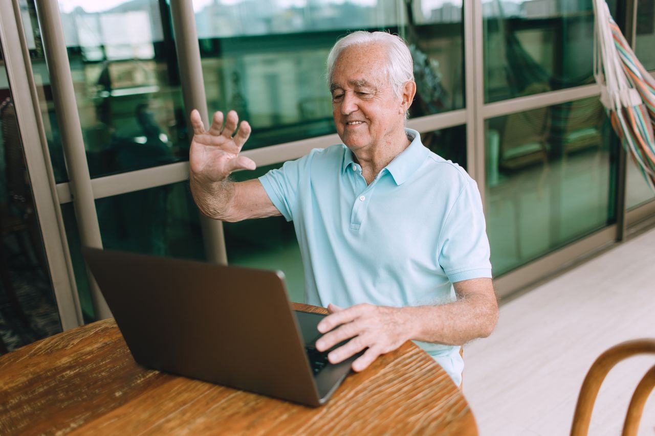 Elderly man using a laptop for a video call indoors, smiling and waving.