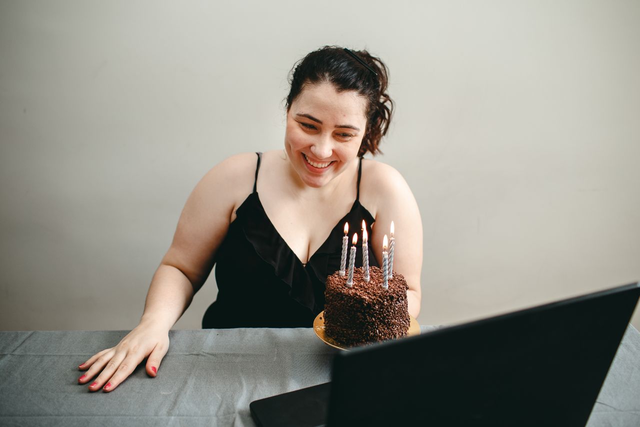 Features Woman smiling at laptop during her virtual birthday celebration with a chocolate cake.