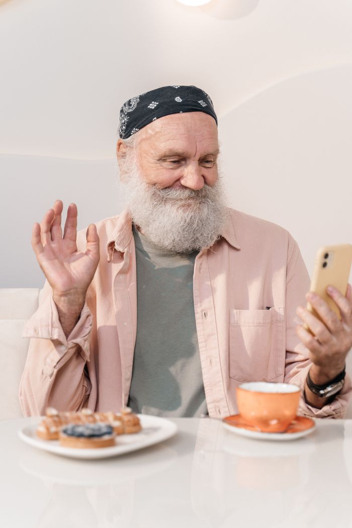 Features Senior man with beard having a virtual chat in a café while enjoying coffee and snacks.